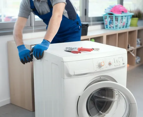 Técnico en overol junto a una lavadora con la puerta abierta y herramientas en la parte superior, preparándose para mostrar cómo reparar el switch de una lavadora Whirlpool.
